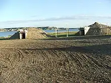 Two dirt-covered earth lodges, with a wooden platform between them and a lake in the background