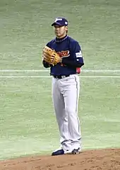 A young Japanese man wearing a dark blue and grey Japan national baseball uniform stands on a pitching mound.