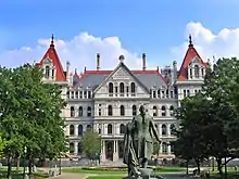 A metallic statue of a man with a short ponytail in his hair, seen from behind, wearing a long coat and holding clothes in his left arm and a walking stick in his right. He is facing a tall stone building in the distance with red peaked roofs. On either side of the image is a row of trees