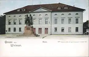 Picture postcard of a large, three-storey building. There is a city square in front of the building. About 10 meters in front of the entrance to the building there is a large bronze statue of two men; the statue is on top of a stone pedestal. The postcard has a white band at its bottom with the printed phrases "Grüss aus Weimar", "Theater", and "499 Verlag von Zedlar und Vogel, Kunstanstalt, Darmstadt 1899."