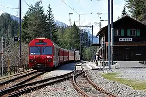 Two-story building with gabled roof next to double-track railway line and platforms; a red train is on the far track