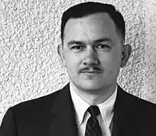 Shoulder-high portrait of a man in his mid thirties with small mustache wearing a black suit with a narrow dark striped tie