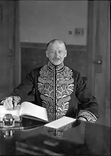 A balding, gray-mustachioed man sitting at a desk with his right hand with a pen on the desk; the man is looking towards the camera and is wearing an elaborately gold-braided chestpiece; all images in this article are black-and-white unless otherwise stated.