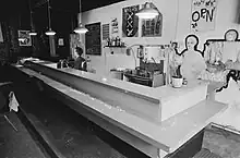 Black and white photograph of an empty bar with sink and beer taps and posters on the wall behind