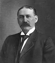 A black-haired man with a handlebar mustache wearing a suit with a necktie and wing collar.