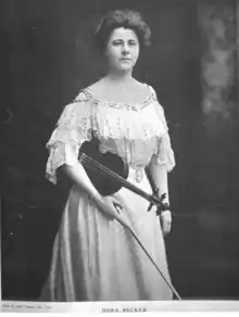 A white woman, standing, wearing a white lacy gown with bare shoulders; she is holding a violin under her right arm, and a bow in her right hand. The background is dark.