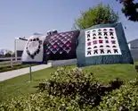  Three examples of Lancaster Amish Quilts shown outside hanging on a line in Lancaster, Pennsylvania. The patterns on these quilts are as follows: A heart made of a wreath of flowers on a white background with a blue block border (Left); Blue, Red, Purple, and green chevrons on a burgundy background (Middle); Depictions of Amish outfits including work shirts and suspenders, hats, pants, and hair coverings on a white background with a teal block border.