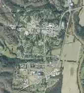 A top-down aerial view of a small, rural town. A river flanked by fields runs along the right side of the frame, while the rest of the town is surrounded by forest.